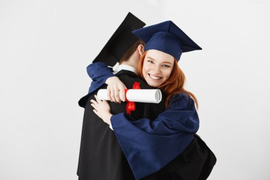Dwóch absolwentów obejmujących się na białym tle. Dziewczyna trzyma dyplom, uśmiechając się, patrząc w aparat.