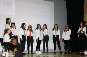 na zdjęciu grupa młodzieży śpiewa piosenkę. wszystkie uczennice ubrane są w czarne spodnie, białe koszule, kolorowe krawaty i okulary przeciwsłoneczne.