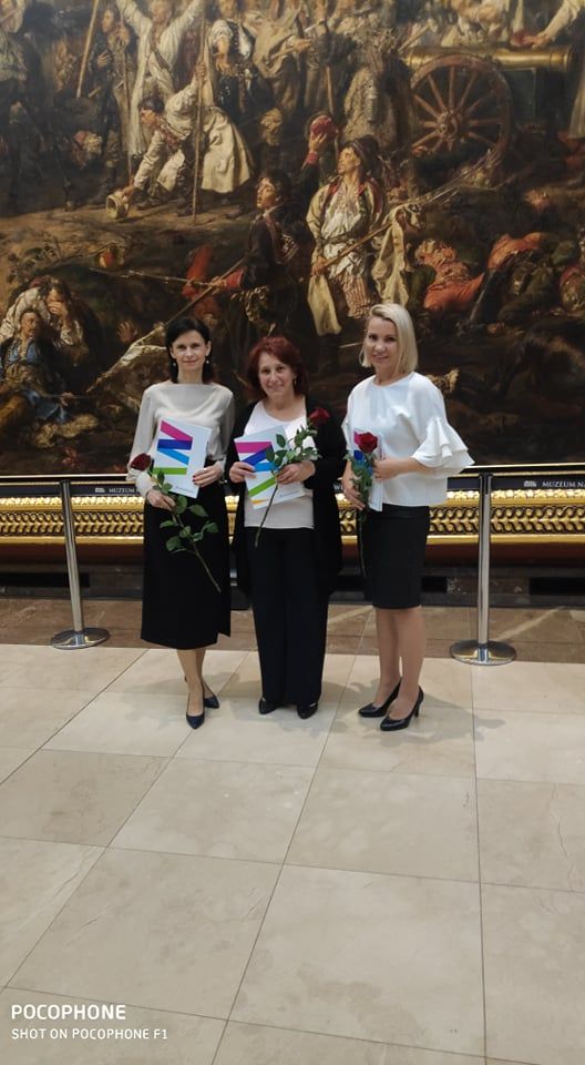 na zdjęciu trzy kobiety w galowych strojach, na tle obrazu w galerii sztuki. Kobiety trzymają w ręku róże i dyplomy